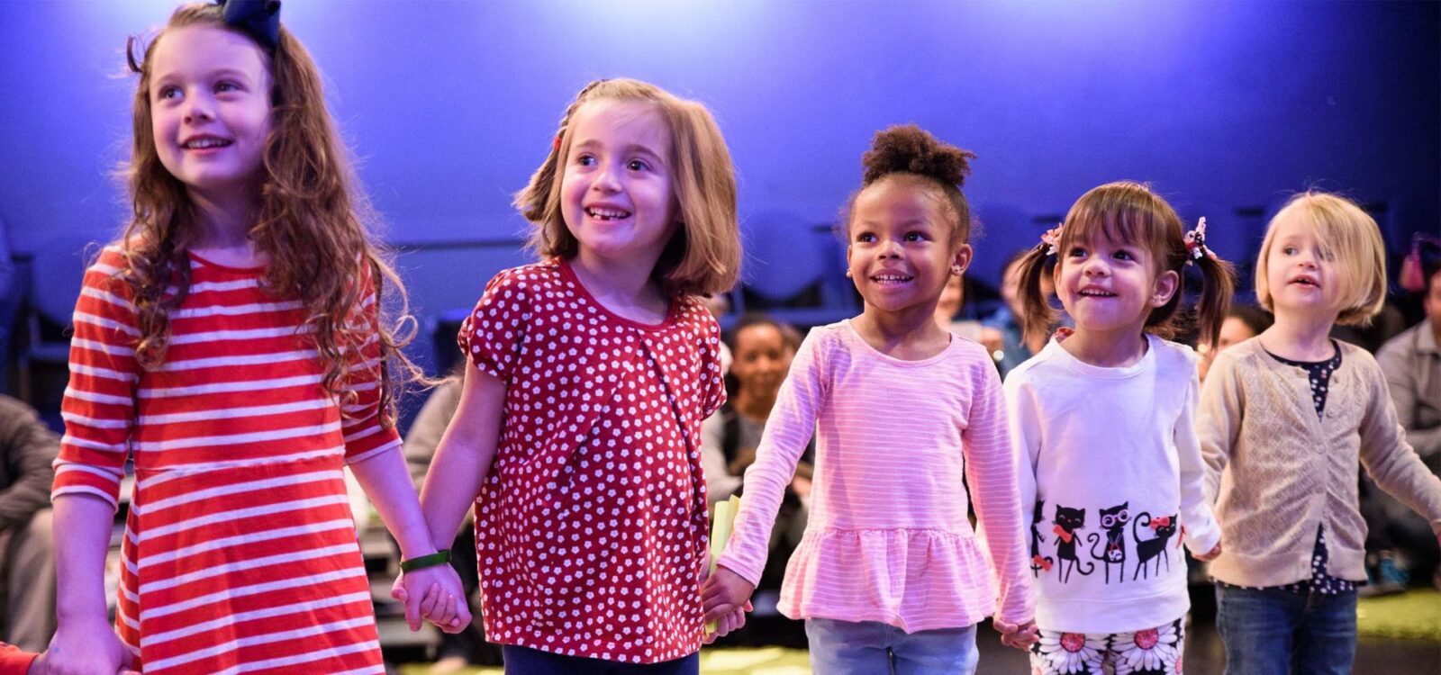 Five young children stand in a row holding hands, smiling, with a blue background and other people sitting behind them.