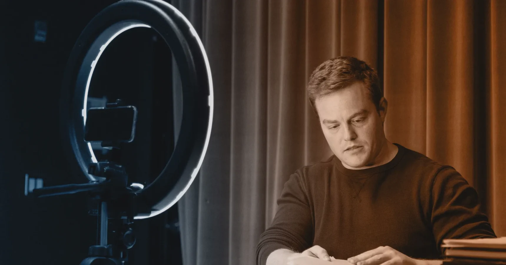 A man sits at a desk in front of a camera and ring light, appearing focused while writing or working on something. Curtains are in the background.