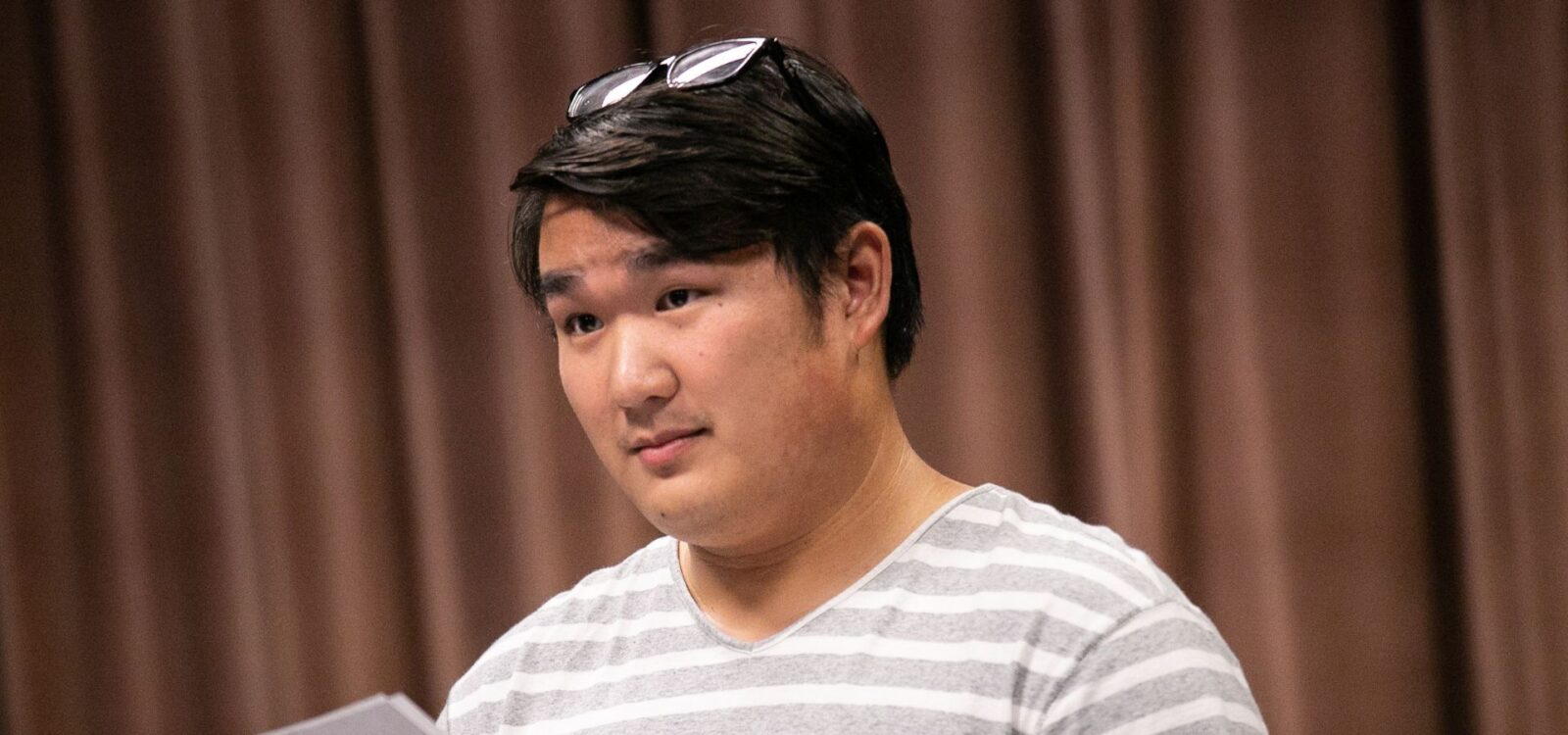 A person in a gray and white striped shirt holds papers, standing in front of brown curtains with sunglasses resting on their head.