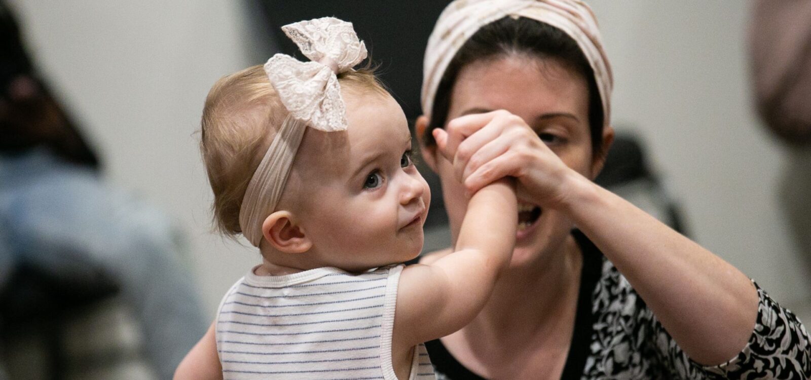 A baby wearing a headband with a bow holds hands with an adult woman, who is partly out of focus, in an indoor setting.