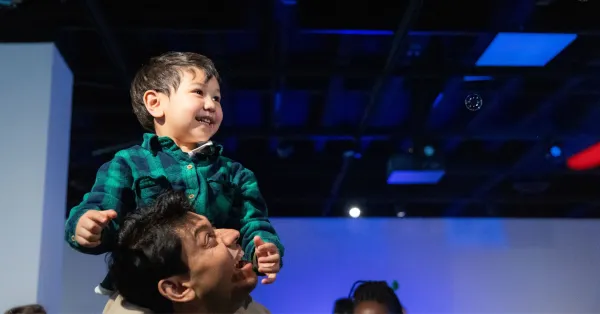 A smiling child sits on an adult’s shoulders, reaching toward a bubble in a dimly lit indoor space.
