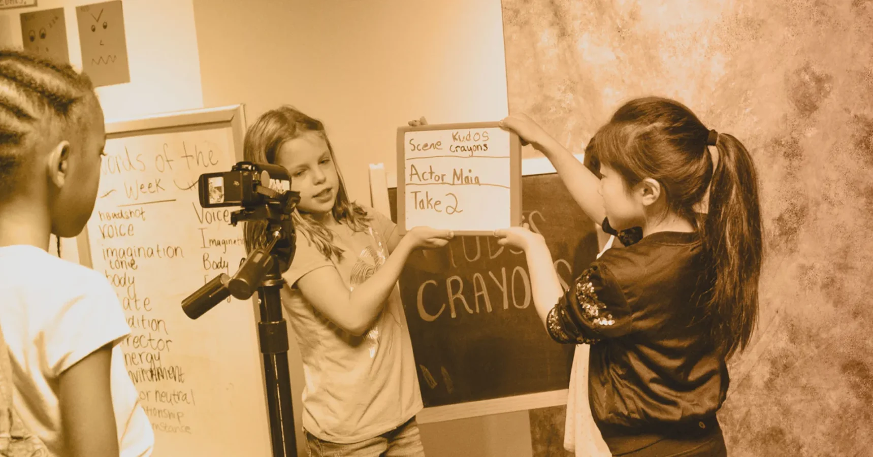 Three children are filming a scene; one operates a camera while two others hold a clapperboard that reads "Actor Mini Take 2." A whiteboard with notes is visible in the background.