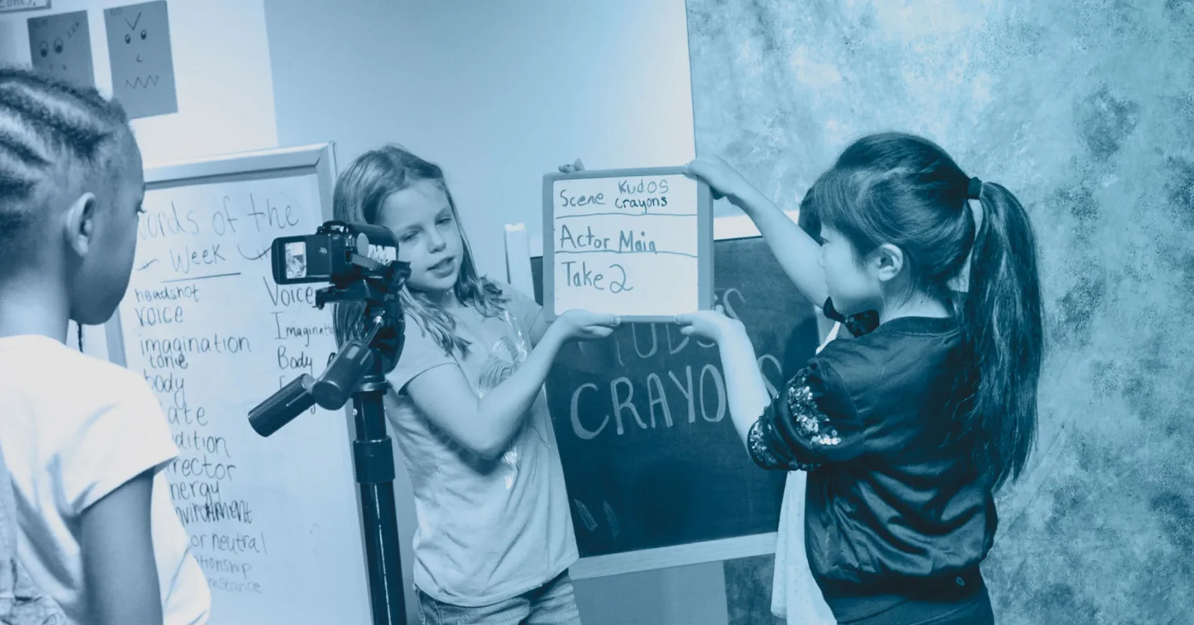 Three girls are filming with a camera; one holds a clapperboard labeled “Scene 1, Take 2,” while another stands nearby, with a whiteboard and chalkboard in the background.