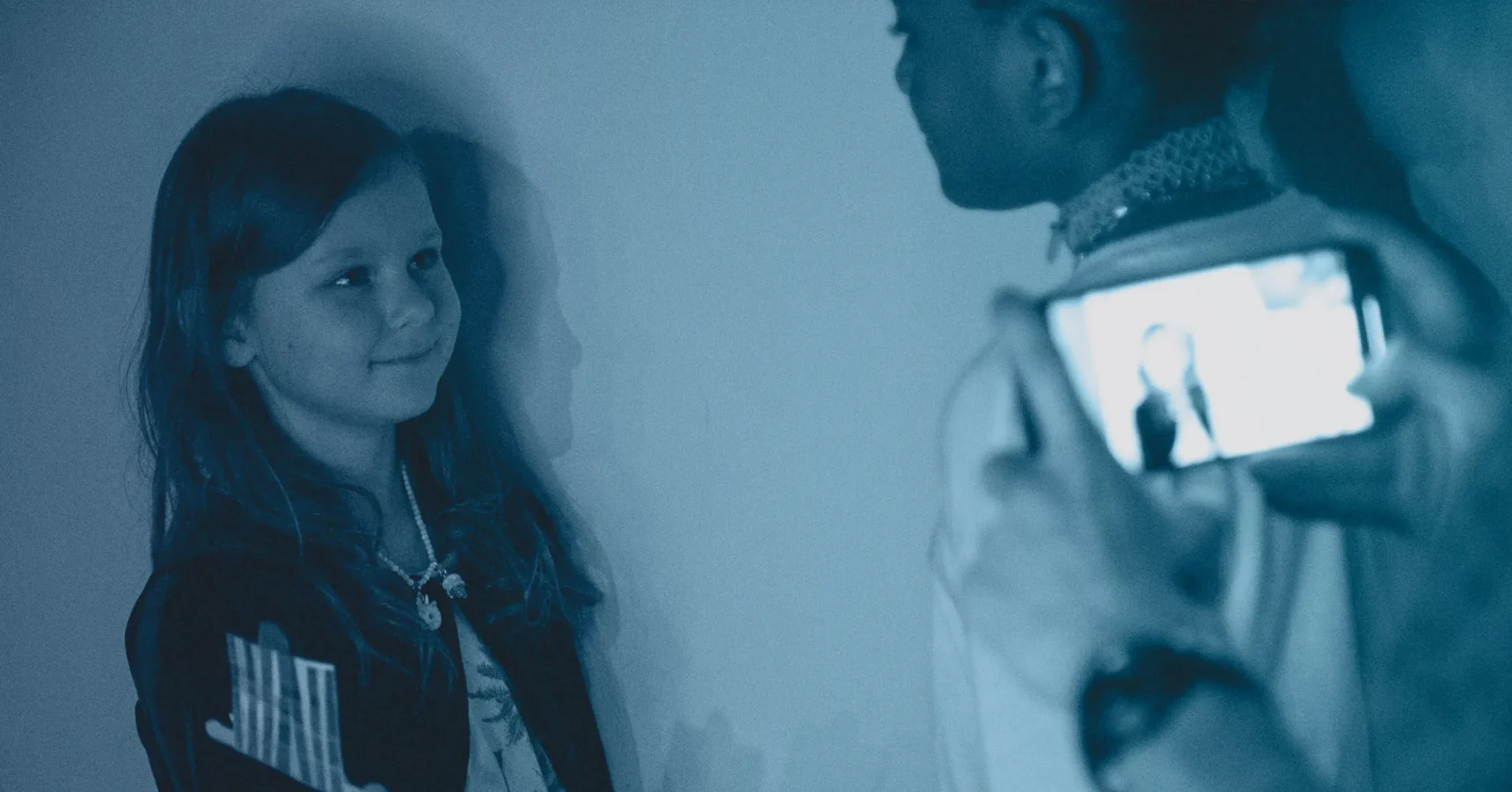 A girl stands against a wall, facing another person as a third person takes a photo of her with a smartphone. The image has a blue tint.