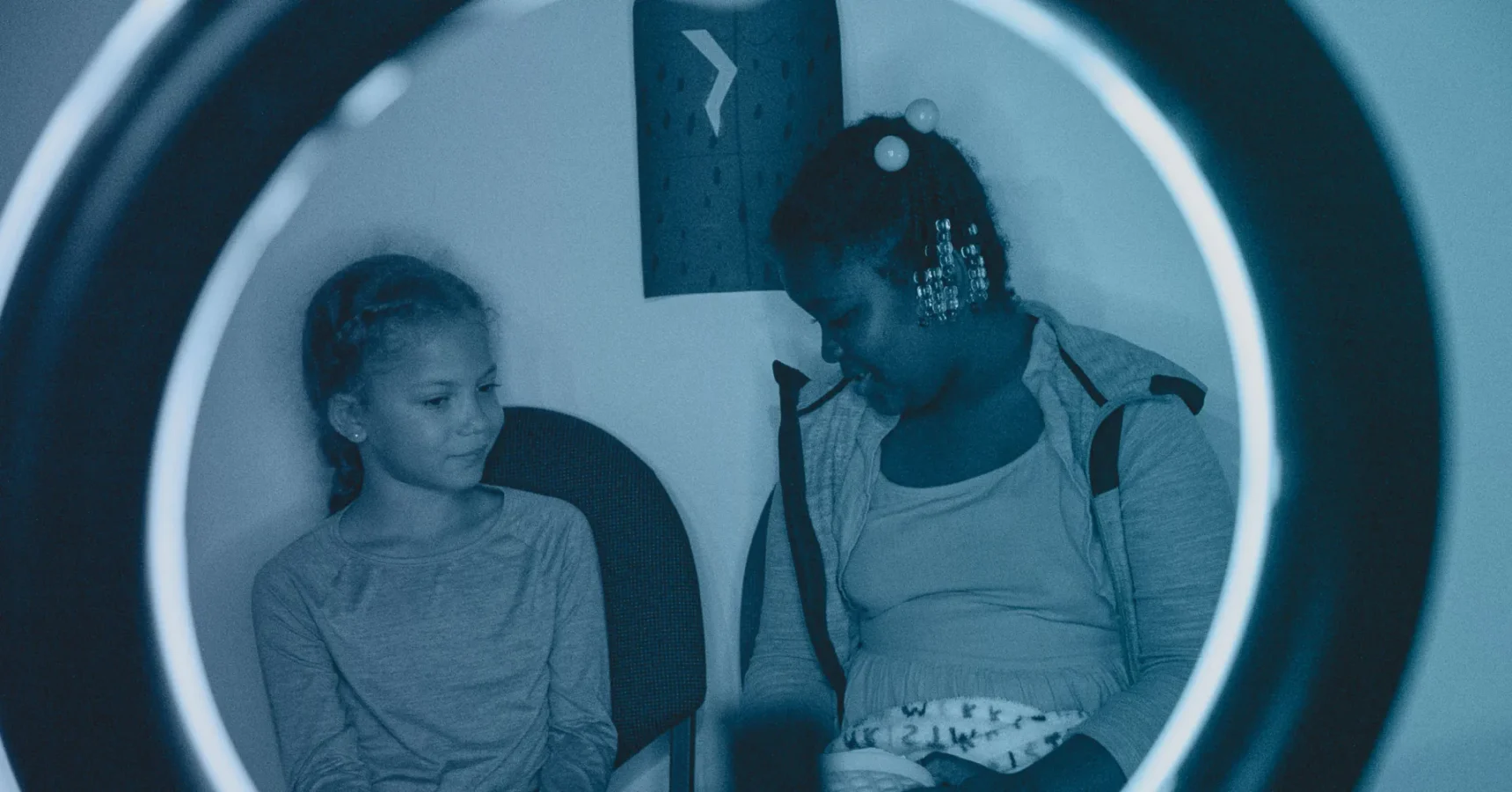Two girls sit next to each other, one looking down and the other looking at her, framed by the circular light of a ring lamp.
