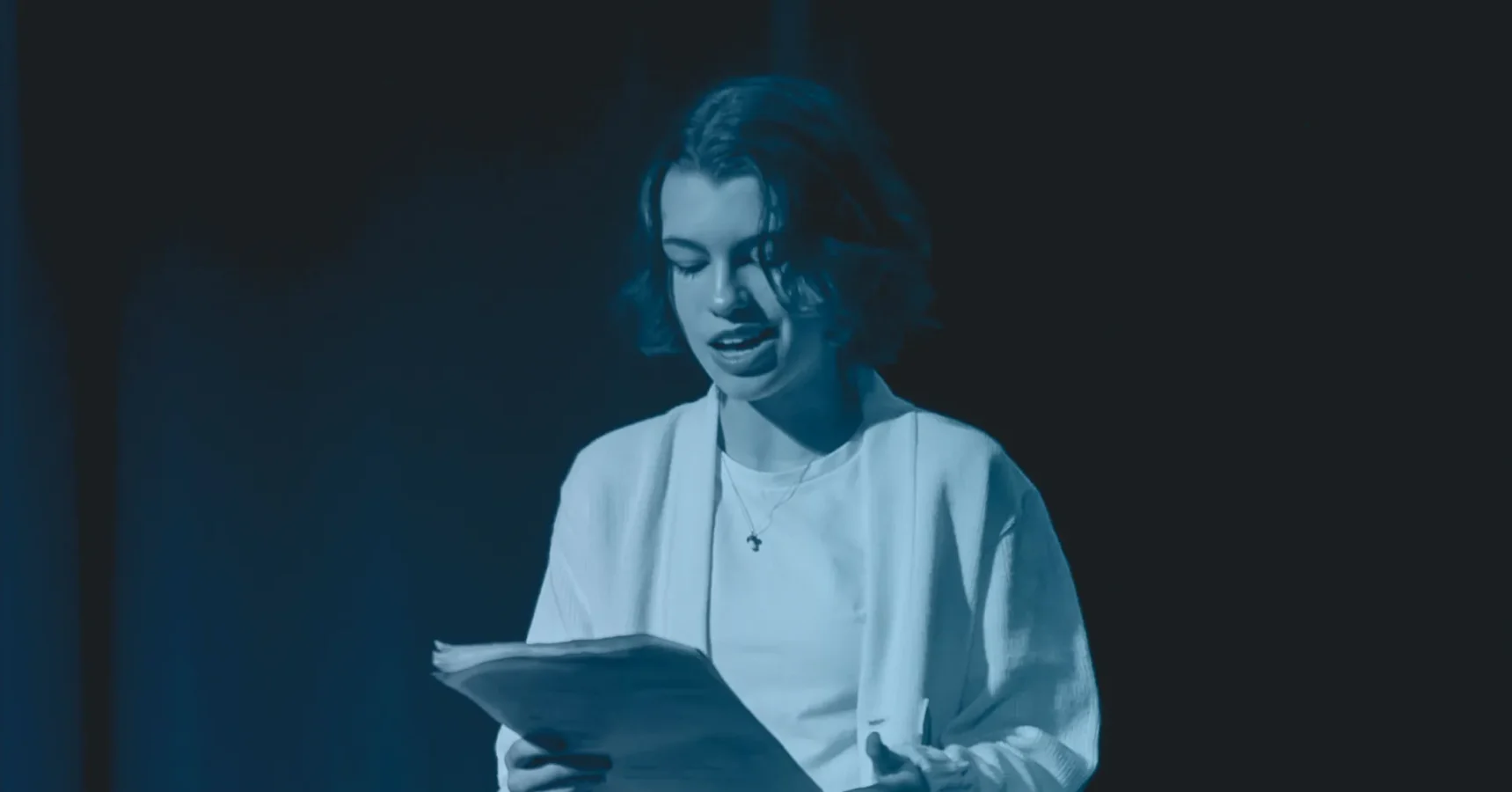 A person with short hair reads from a stack of papers while standing on a dark stage, illuminated by blue lighting.