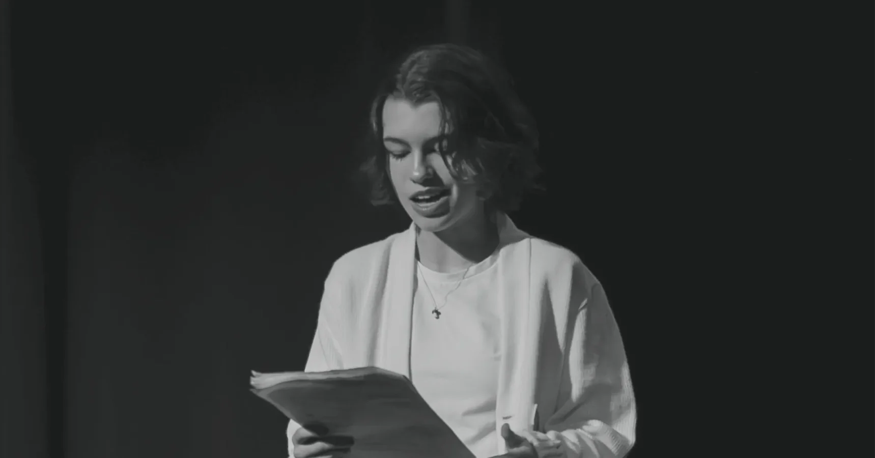 A person with short hair reads from a stack of papers, standing in front of a dark background.