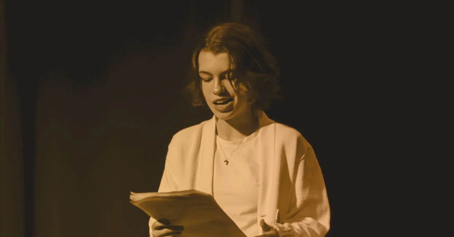 A person stands on stage holding a script, speaking or performing, with a dark background and focused lighting.