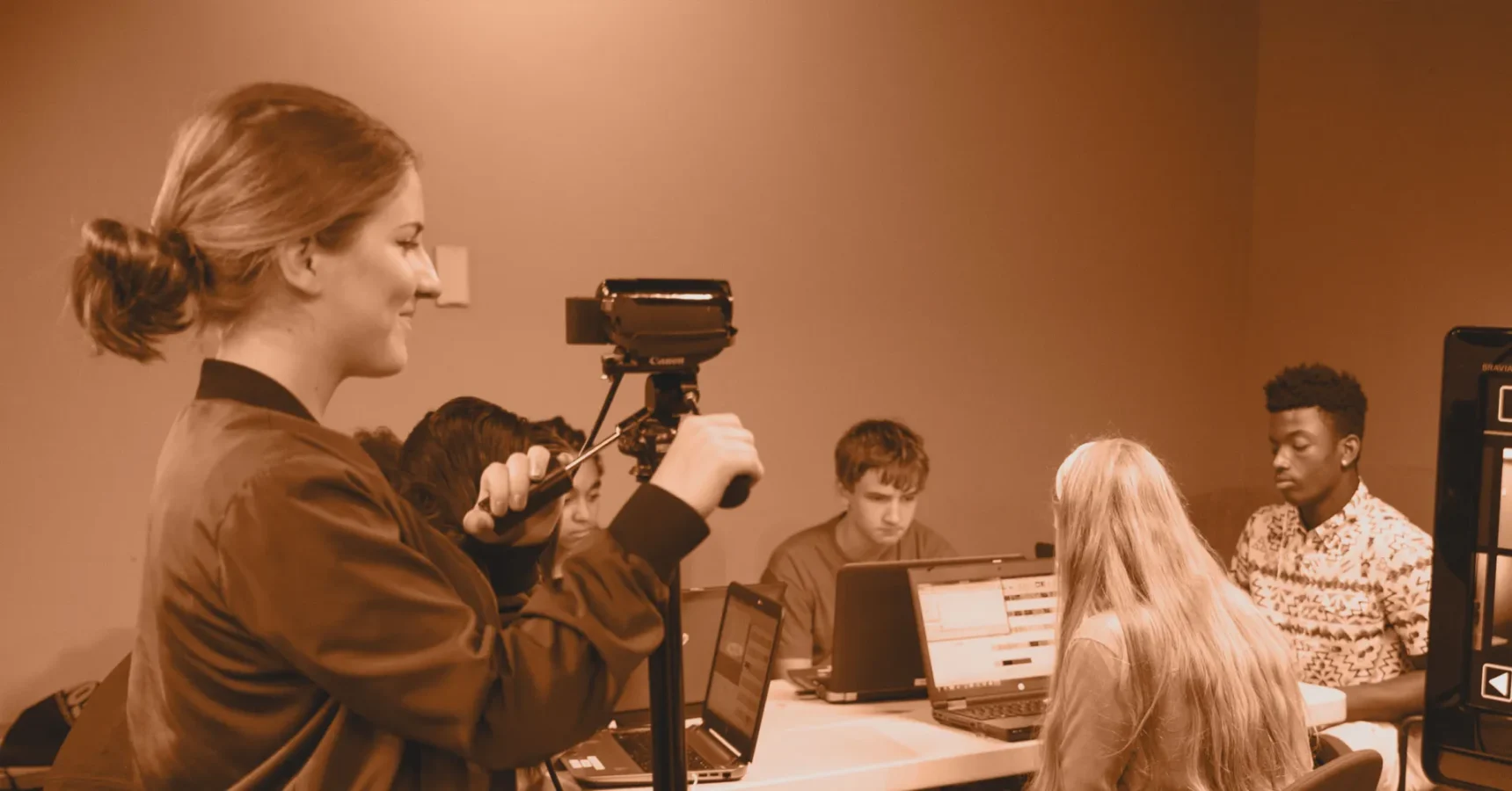 A group of young people work on laptops around a table while one person stands recording video with a camera on a tripod.