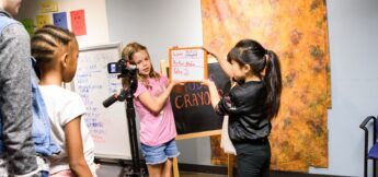 Four children stand in a classroom, two holding a clapperboard in front of a camera on a tripod, preparing to film against an orange and brown backdrop in camera acting camp.