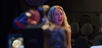 A middle school student with long blonde hair smiles at camera acting camp, speaking in a dimly lit room with empty chairs and camera equipment visible in the foreground.