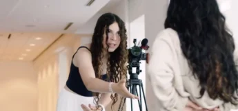 A teen with long curly hair gestures while speaking to another person in front of a camera on a tripod in camera acting camp for high school students.