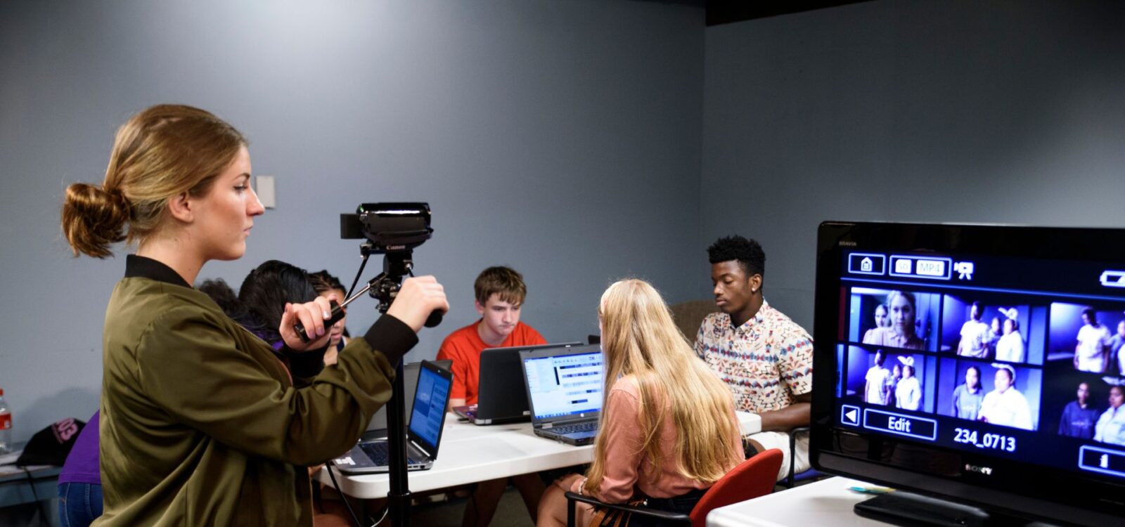 A woman adjusts a video camera while four people work on laptops at a table; a monitor displays images from the camera in a classroom setting.