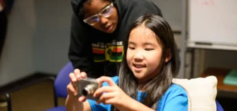 A young girl holds and looks at a digital camera while a teacher stands behind her, observing her actions in Camera Acting: Shorts Camp.