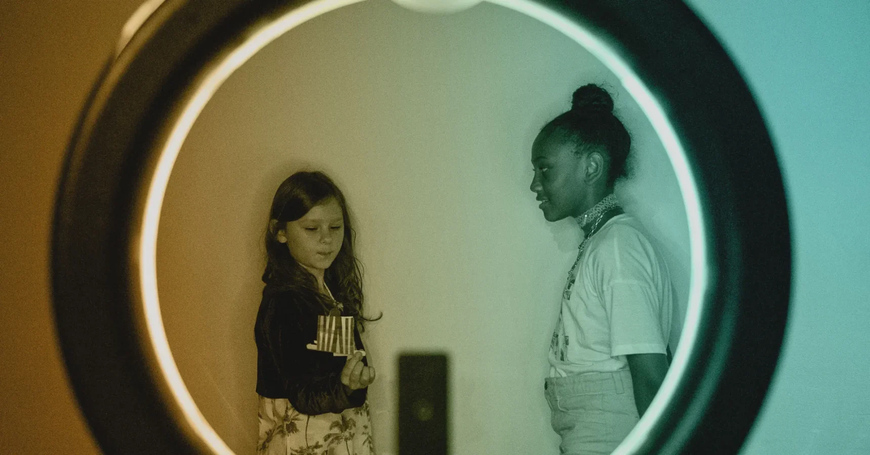 Two girls stand facing each other indoors, framed by a circular ring light in the foreground.