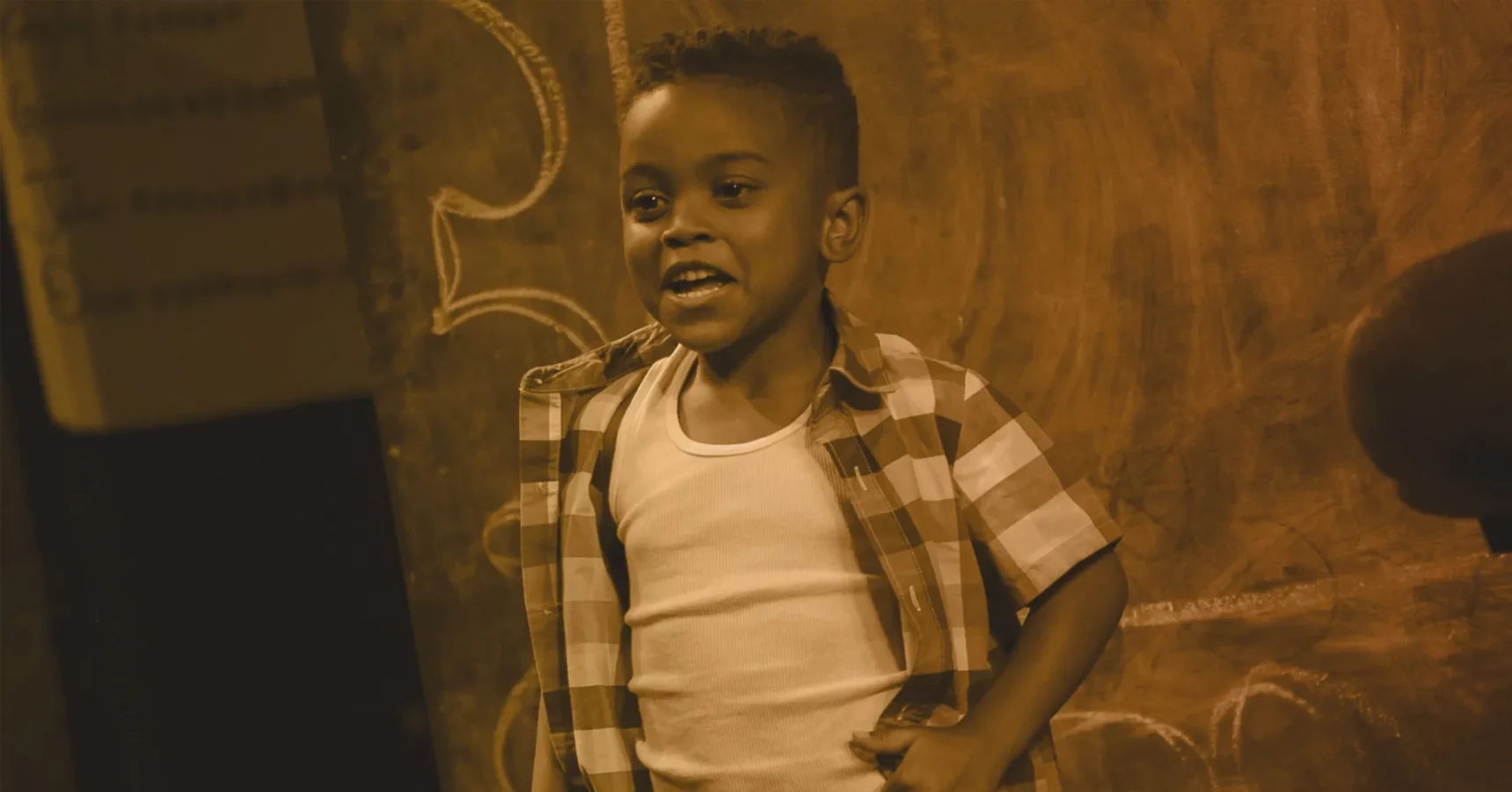 A young boy in a plaid shirt and white undershirt stands in front of a chalkboard, smiling and speaking.