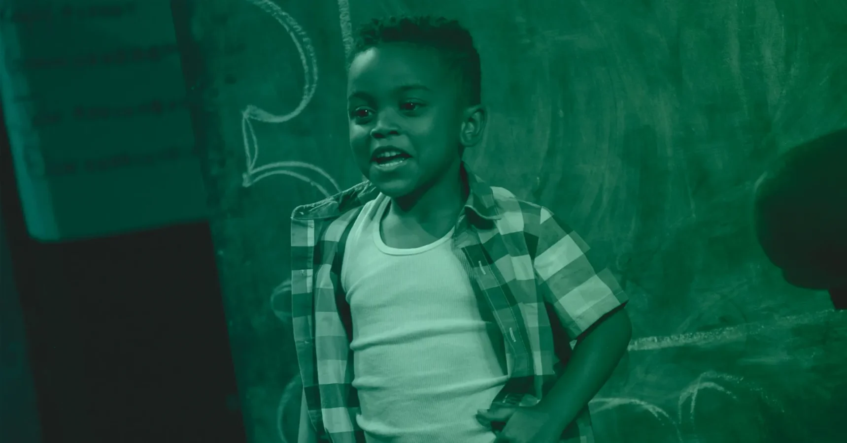 A young boy wearing a plaid shirt and white tank top stands in front of a chalkboard with hand-drawn designs, looking slightly to the side.