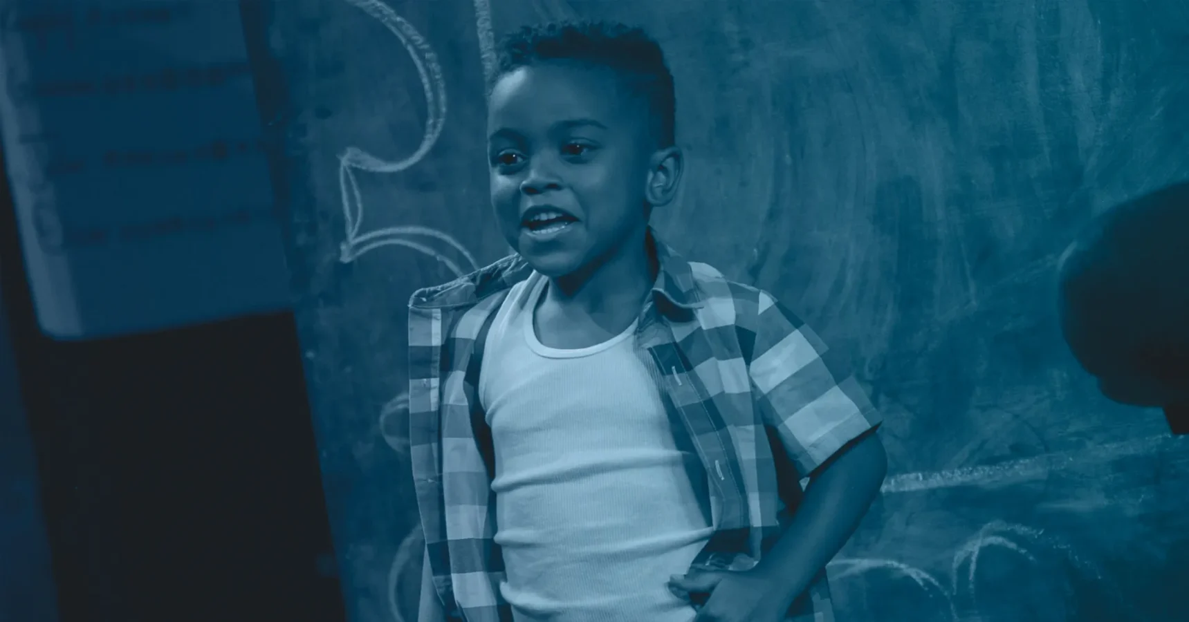 A young boy in a plaid shirt and white tank top stands in front of a chalkboard with drawings, smiling and looking slightly to the side.