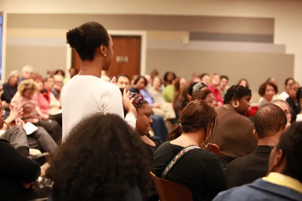 A woman stands and speaks in a crowded room filled with seated people attending a community meeting or seminar.