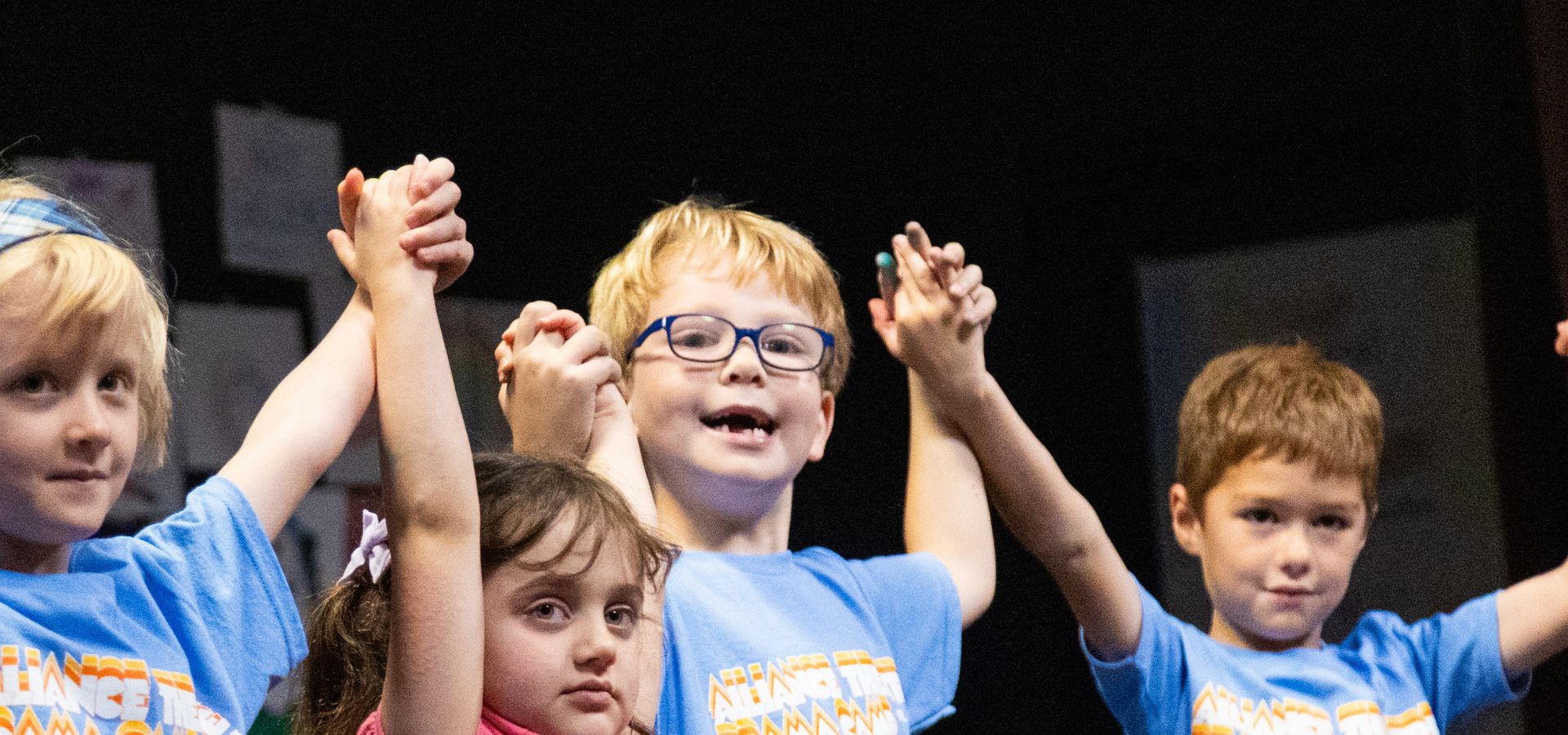 Four young children stand in a row holding hands up, wearing blue shirts in creative drama camp. The child in the center smiles, while others look toward the camera.