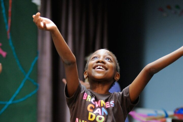 A child in a brown t-shirt raises both arms and smiles joyfully indoors, with other children visible in the background.