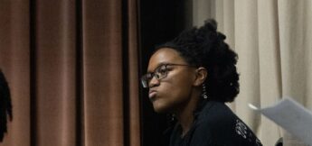 A person wearing glasses and a black shirt looks to the side with a neutral expression, seated indoors near beige and brown curtains.