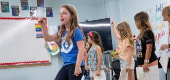 A group of children stand in a classroom, holding papers and raising their hands, with one girl in front appearing to speak or sing in musical theater dance camp.