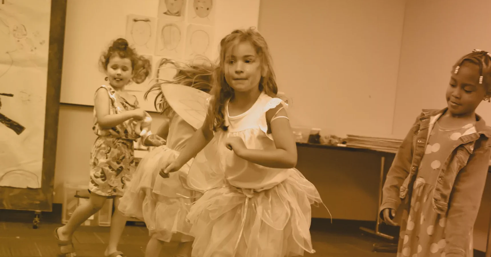 Three young girls in costumes dance in a classroom, while a fourth girl in a polka-dot dress and jacket watches. Drawings are visible on the wall in the background.