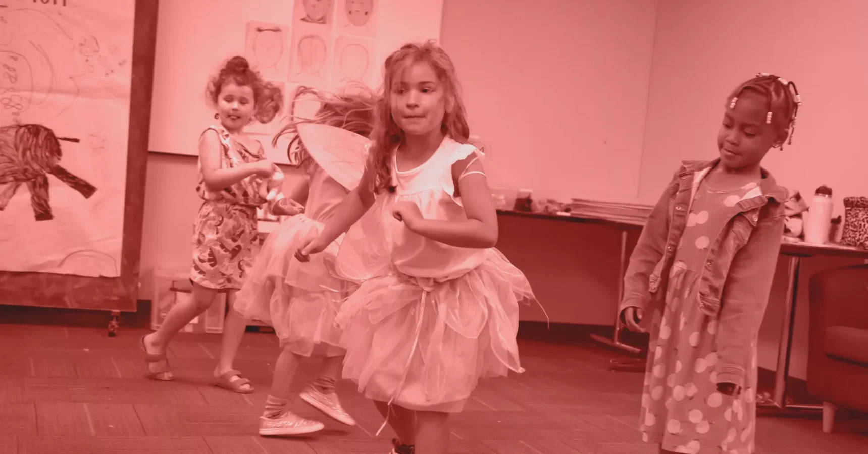 Four young children wearing costumes and dresses dance and play in a classroom decorated with drawings on the walls.