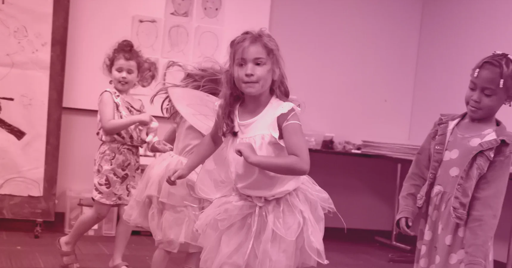 Three young children in costumes, including tutus and wings, play and dance in a classroom decorated with drawings on the wall.