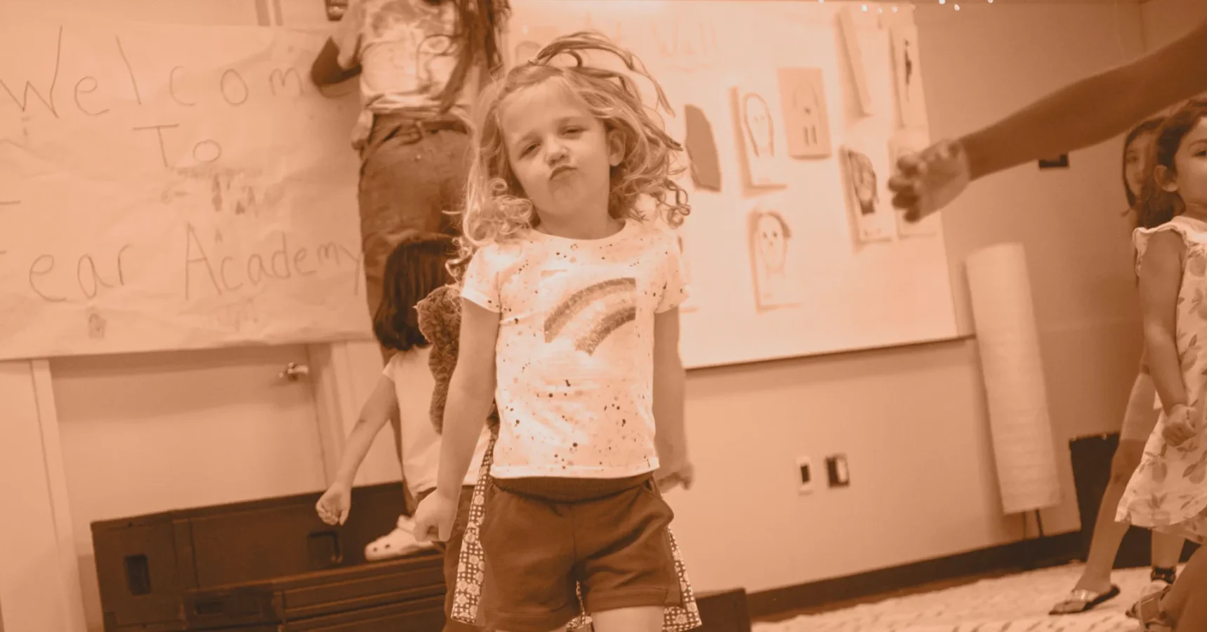 Young girl stands in the center of a classroom with other children, wearing a t-shirt with a rainbow, while art and a welcome sign hang on the walls in the background.