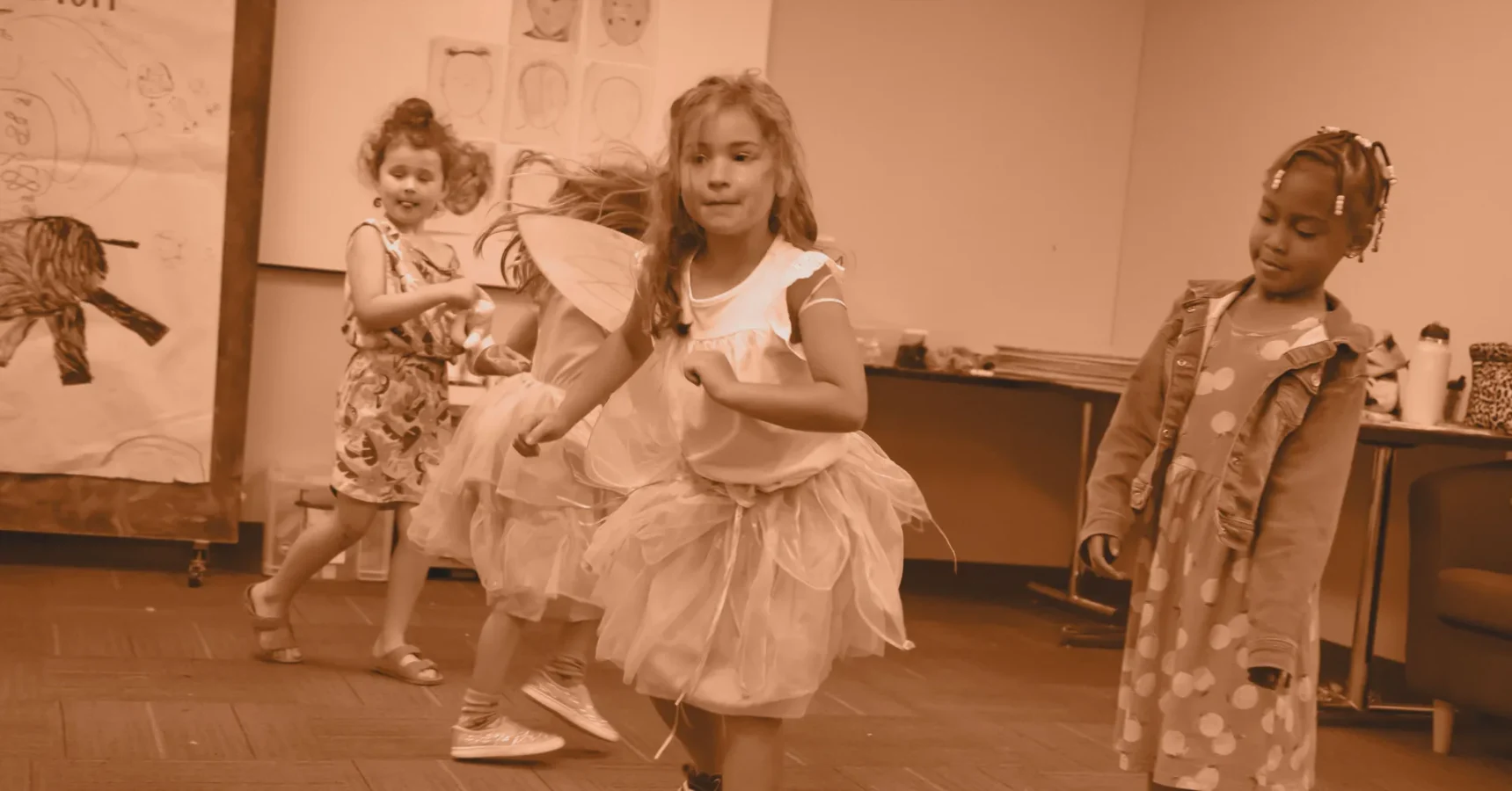 Four young girls wearing costumes dance and play together in a classroom. Drawings are displayed on the wall in the background.