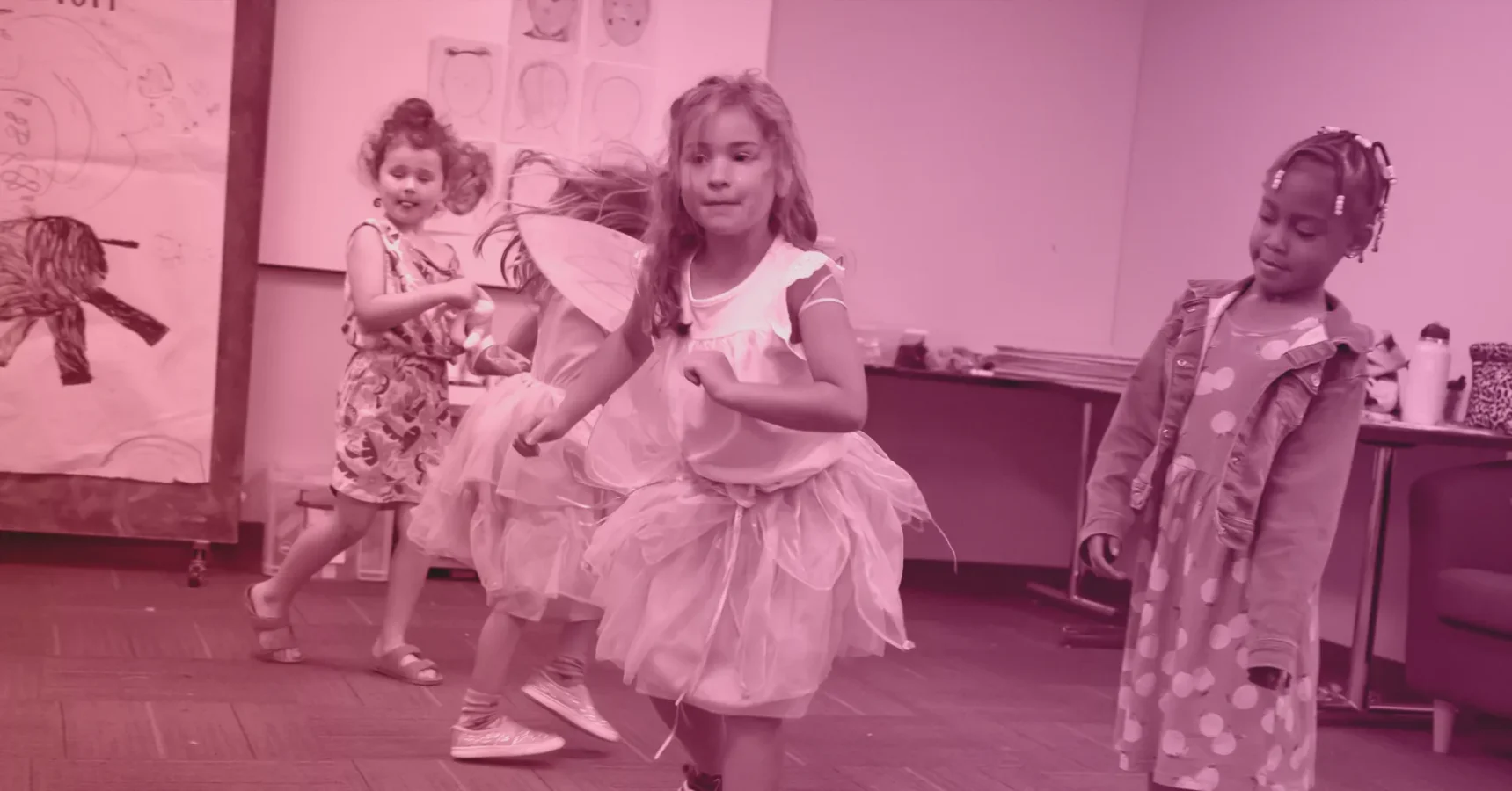 Three young girls in dresses and fairy wings play together indoors, while a fourth girl in a polka-dot coat stands nearby; children's drawings are on the wall behind them.