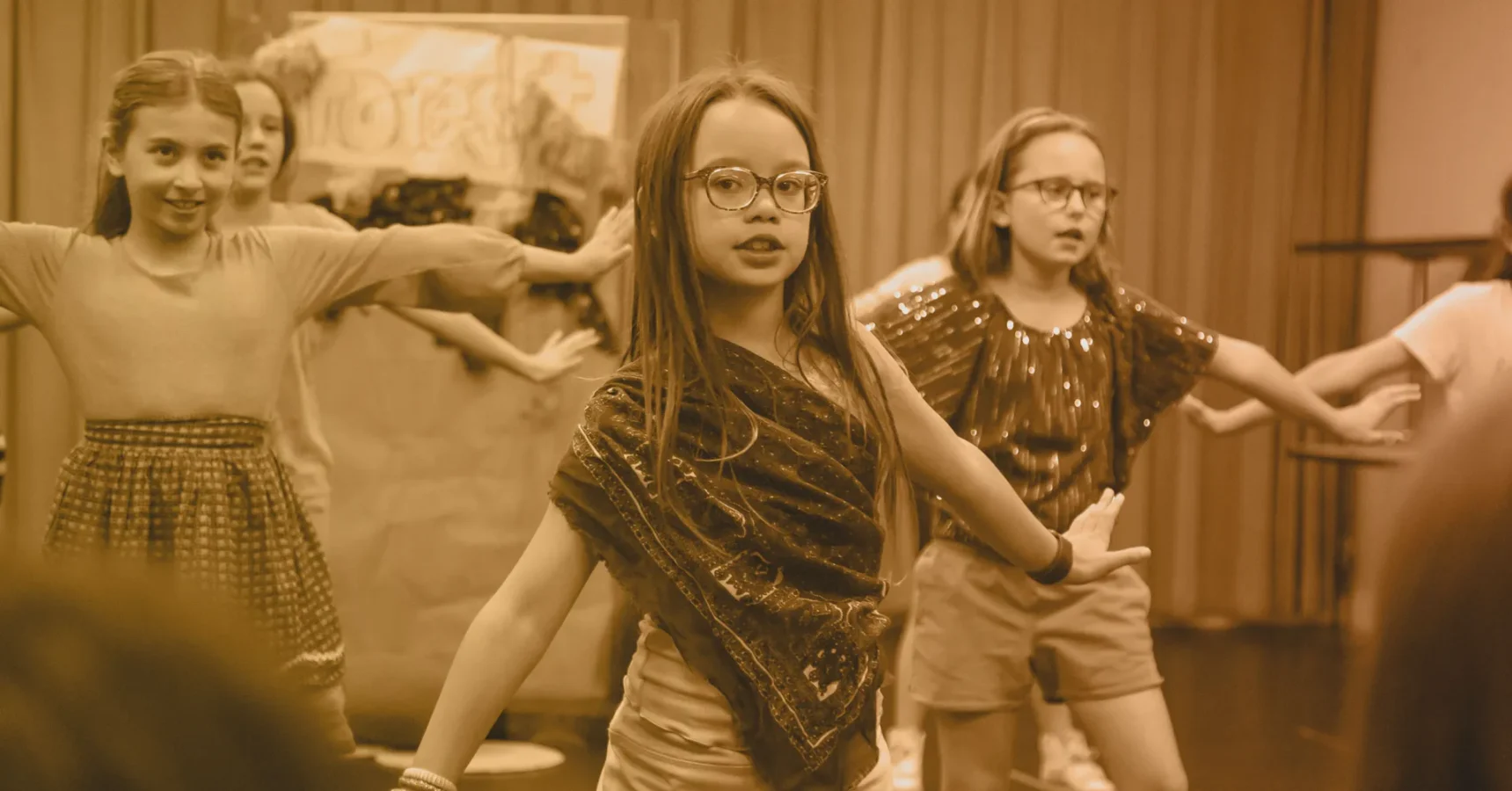 A group of children perform on stage, with one girl in glasses standing in front and others posing with arms outstretched in the background.