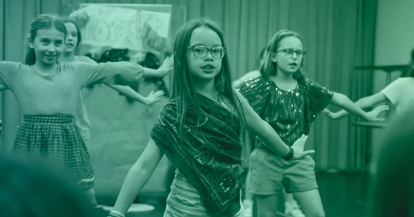 Three young girls perform a choreographed dance on stage, wearing casual clothes and glasses, with a decorated backdrop behind them.