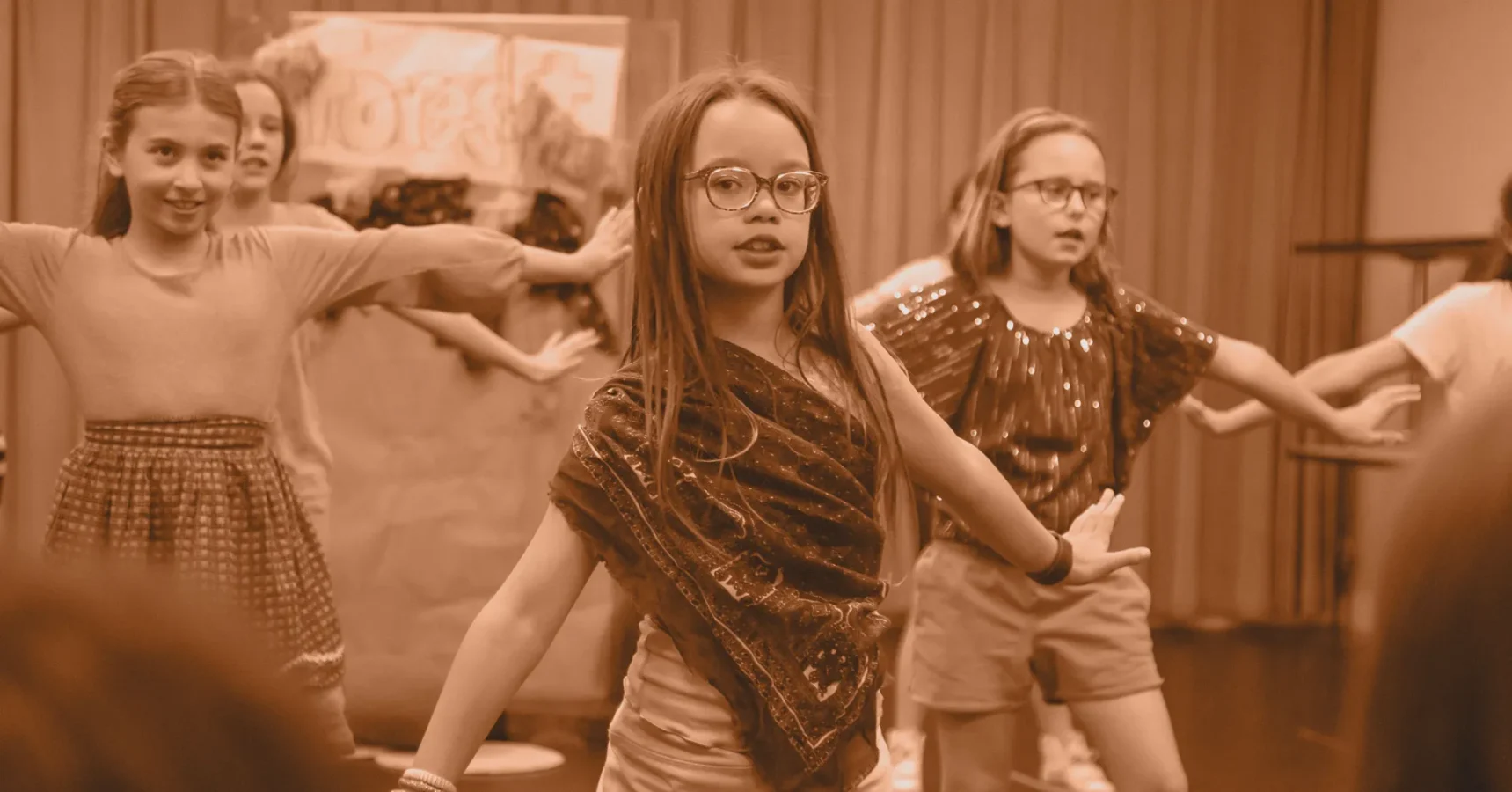 Several children stand with arms outstretched, performing a group dance or play on stage in front of an audience.