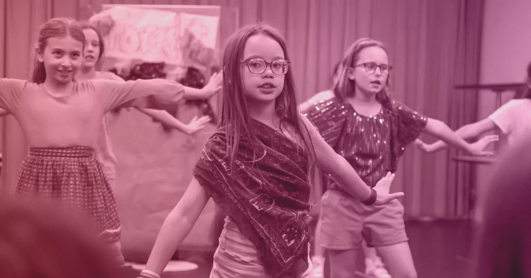 A group of children perform a choreographed dance on stage, with arms extended, in front of a curtain and backdrop.