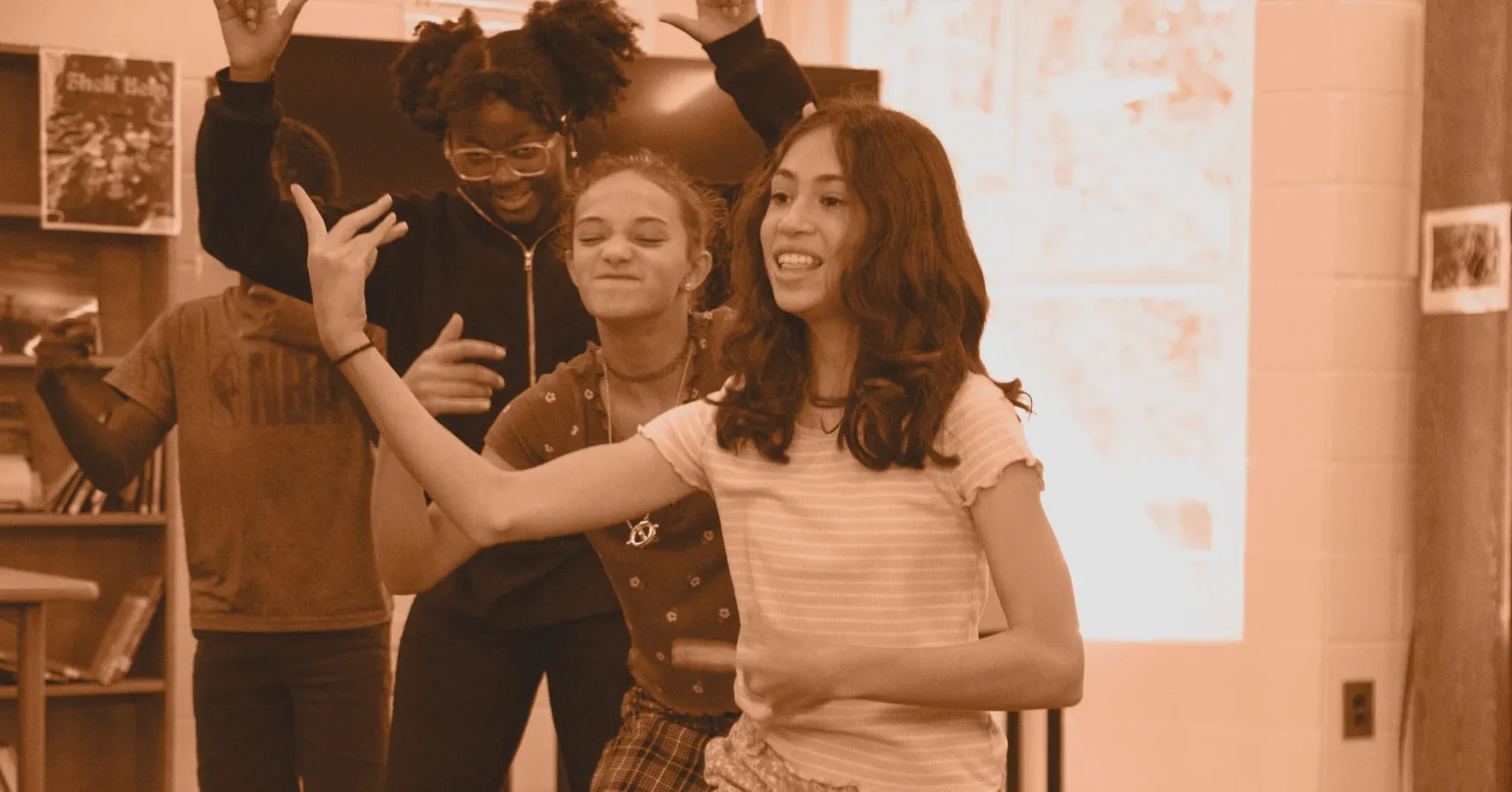 Four teenagers stand indoors, smiling and posing with raised hands in a well-lit room with bookshelves and posters in the background.