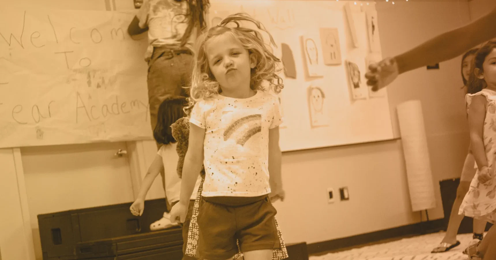 Young girl with curly hair poses with a playful expression in a classroom setting; children and artwork are visible in the background.