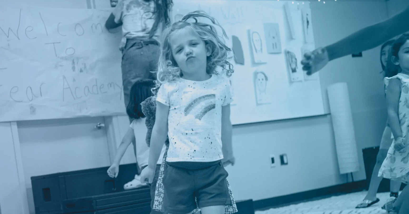 A young girl stands confidently in a classroom with other children in the background, colorful artwork and a welcome sign on the walls.