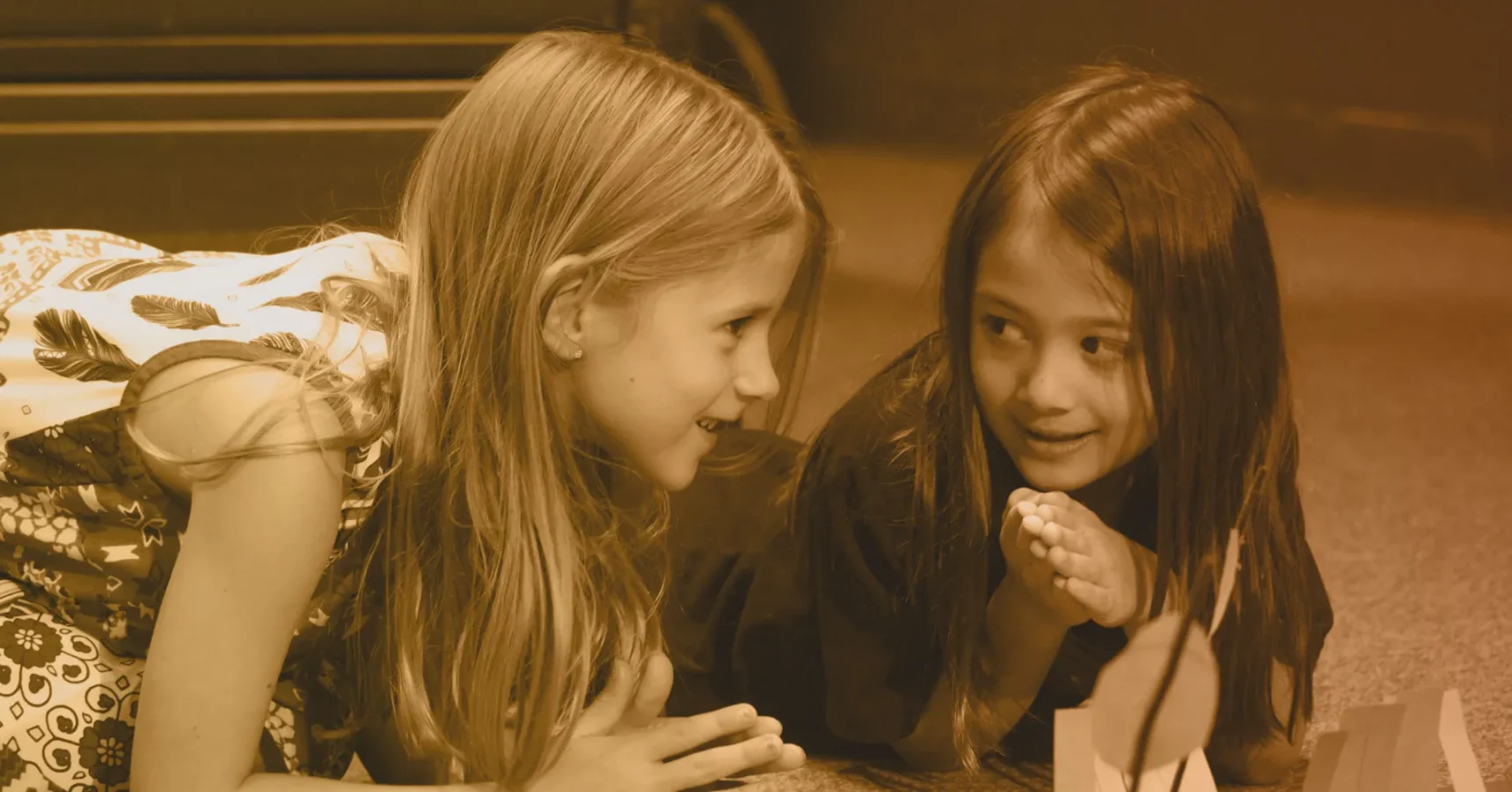 Two young girls lie on the floor facing each other, smiling and talking, with hands near their faces.