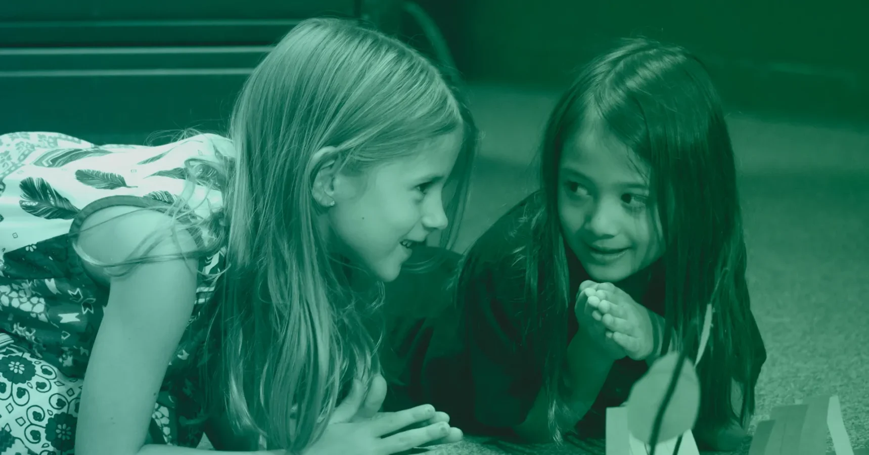 Two young girls lie on the floor facing each other, smiling and talking, with hands clasped near their faces.