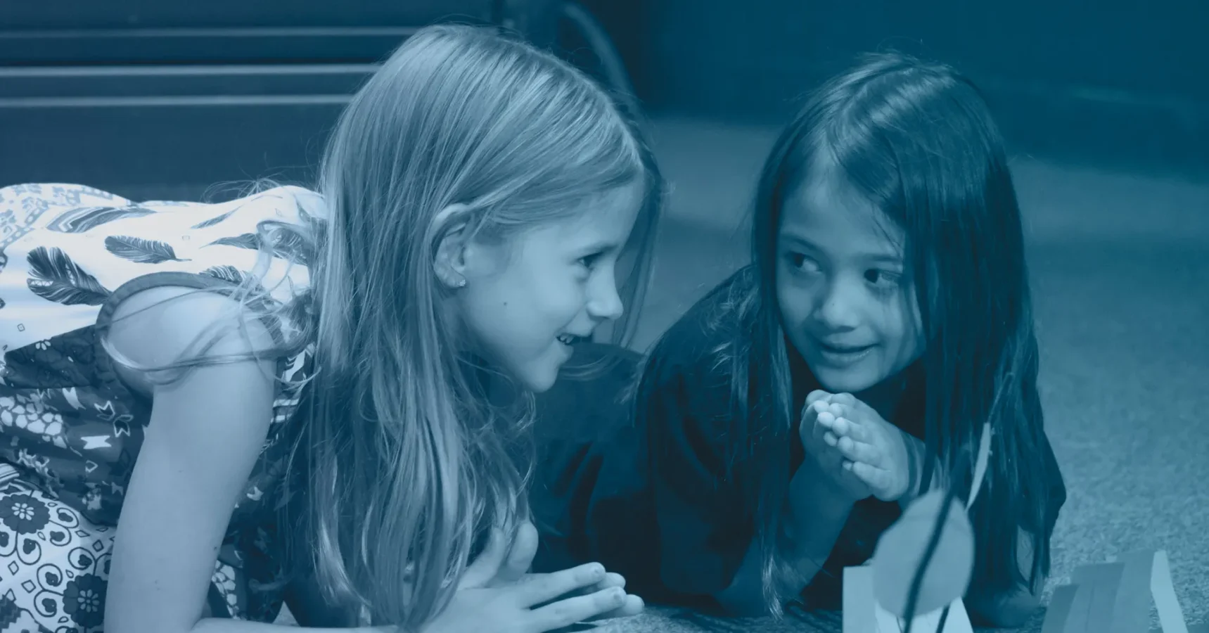 Two young girls lie on the floor facing each other, smiling and talking, with hands clasped in front of them near a small paper craft.