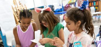 Three young children in a classroom work on crafts, wearing paper hats and headbands, focused on their creative projects at a table in design camp.