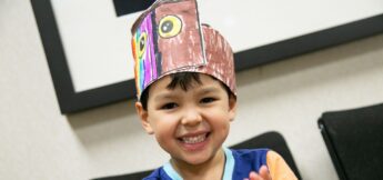 A young child smiles while wearing a handmade paper owl hat with colorful drawings in design camp, sitting indoors in front of a picture frame.