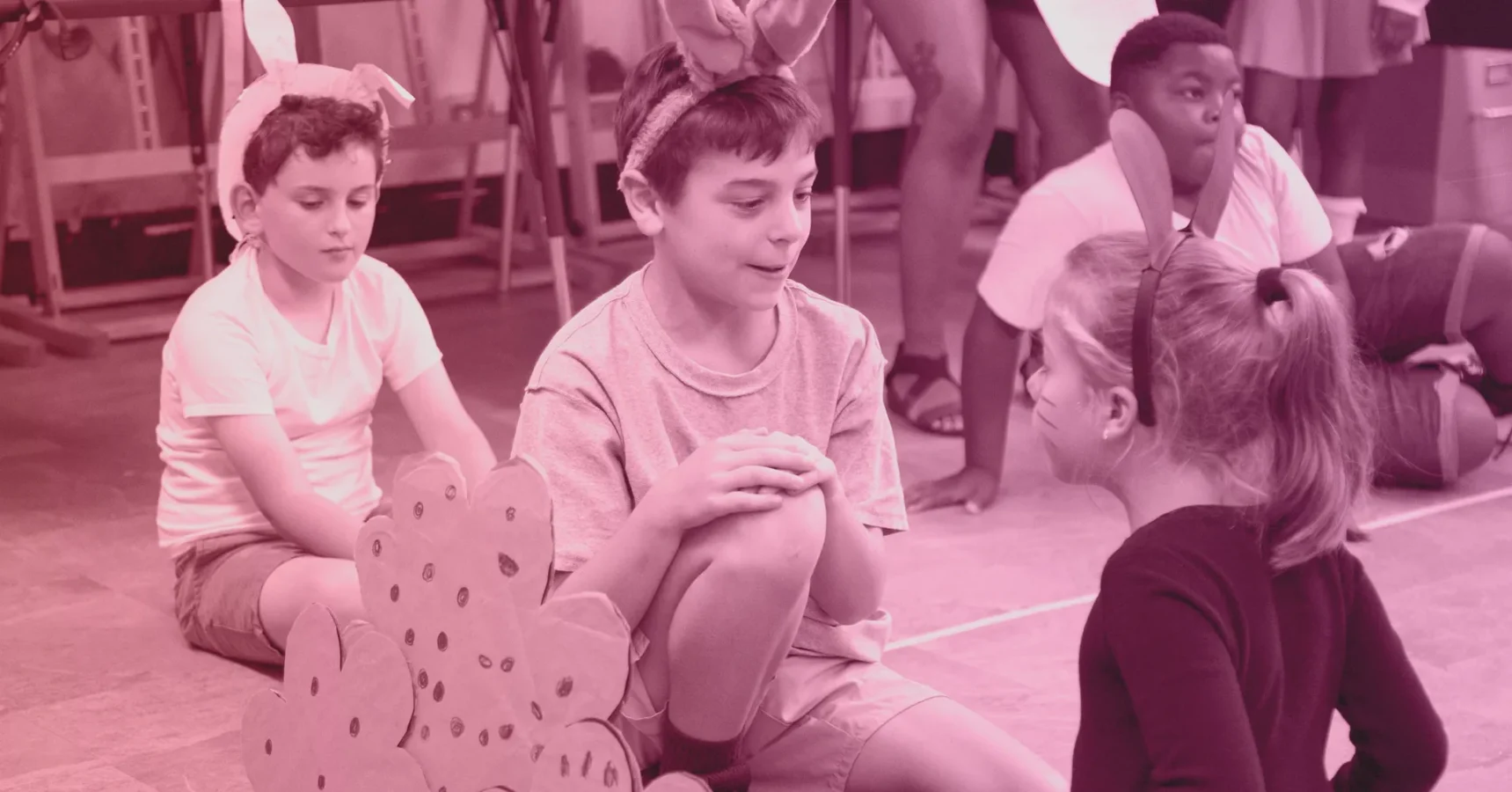 Three children wearing bunny ear headbands sit on the floor, engaged in conversation, with other children and stage props in the background.