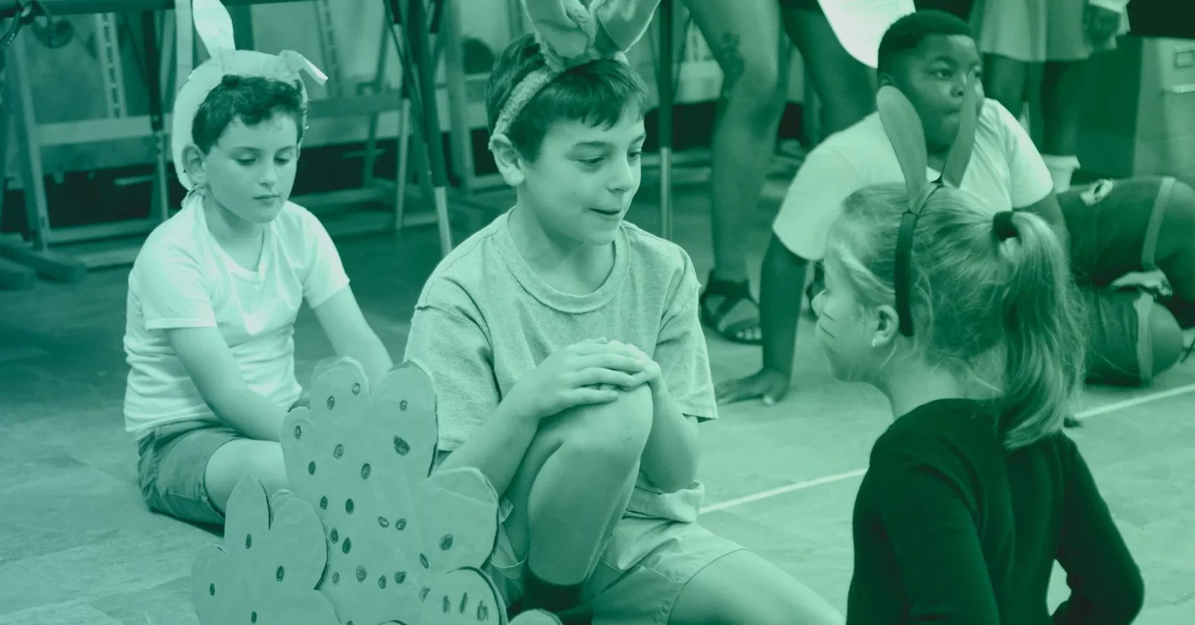 Three children wearing animal ears sit on the floor, talking, with other children and cardboard props visible in the background.