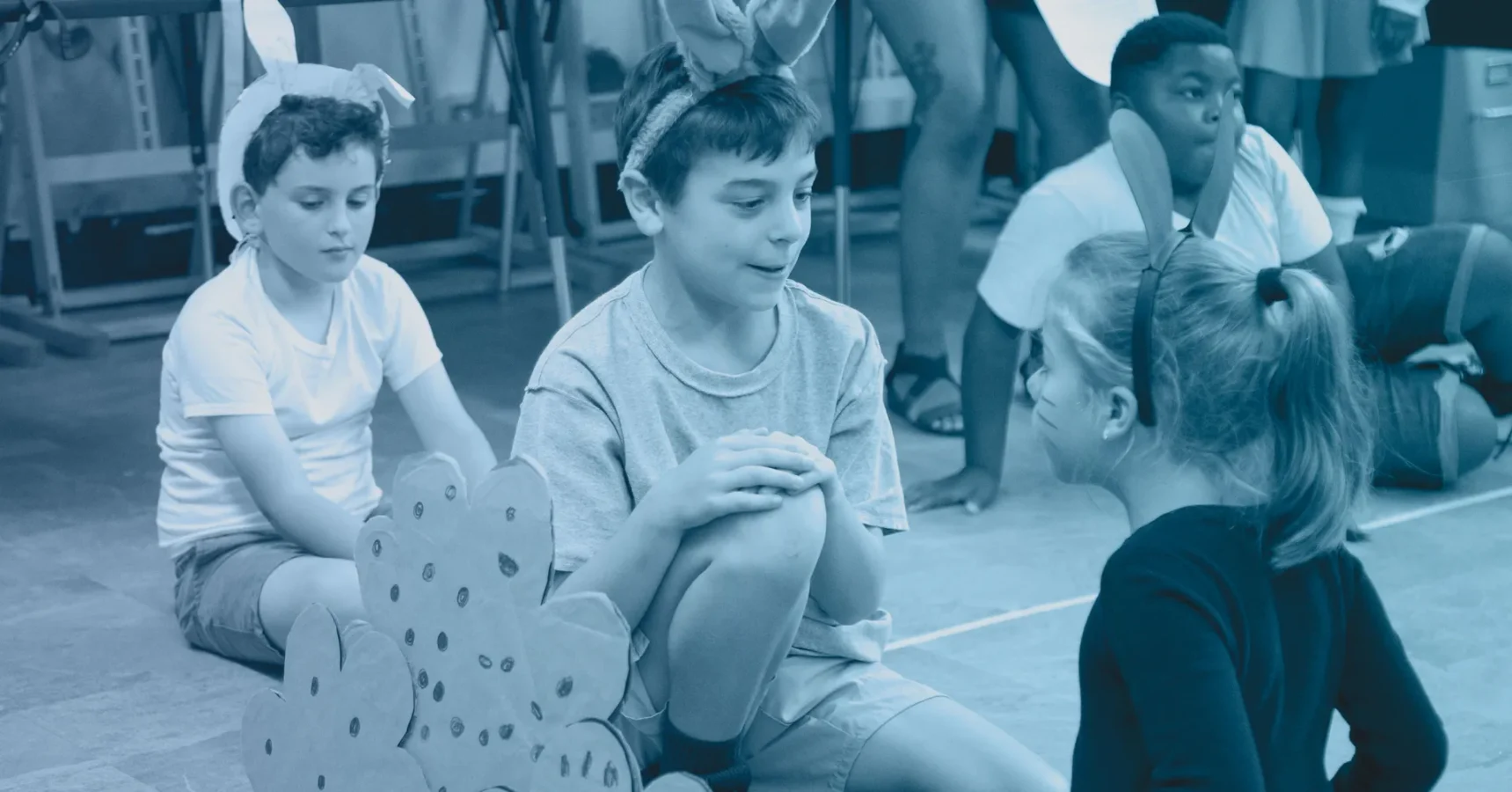 Children wearing bunny ear headbands sit on the floor in a classroom, participating in an activity or play.