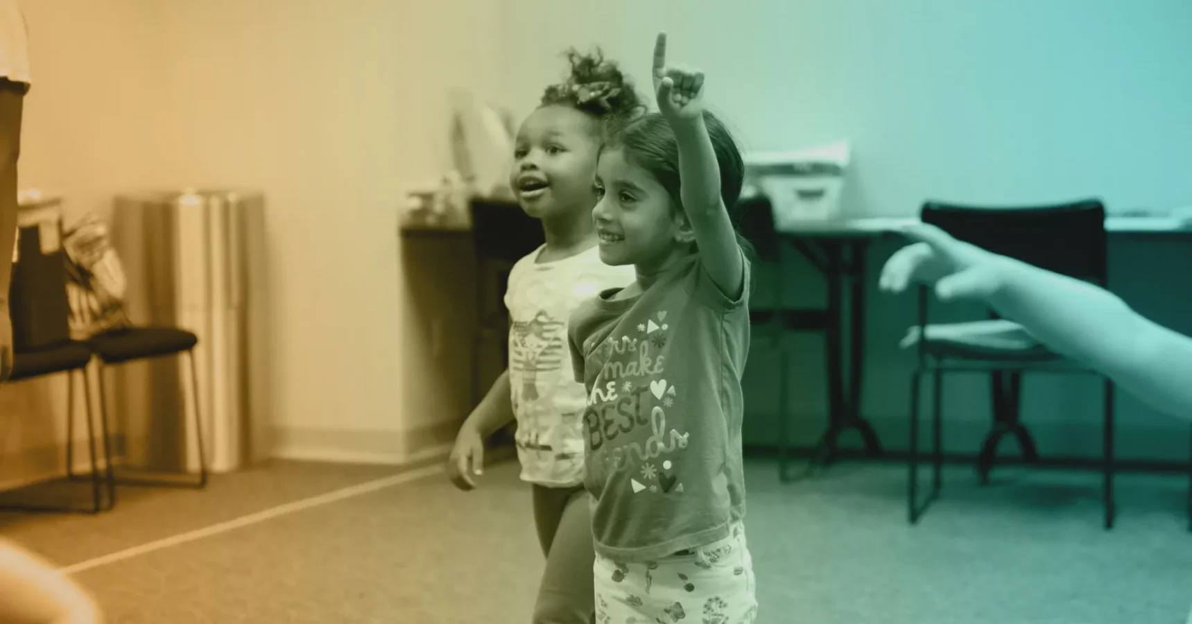 Two young children stand indoors; one is smiling and raising a hand while the other looks on. Tables and chairs are in the background.