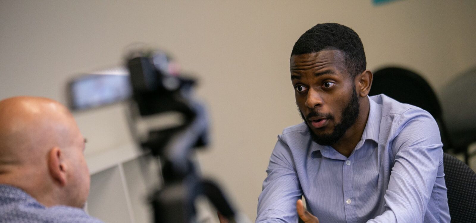 Two men sit across from each other at a table having a conversation, with a camera set up between them recording the discussion.